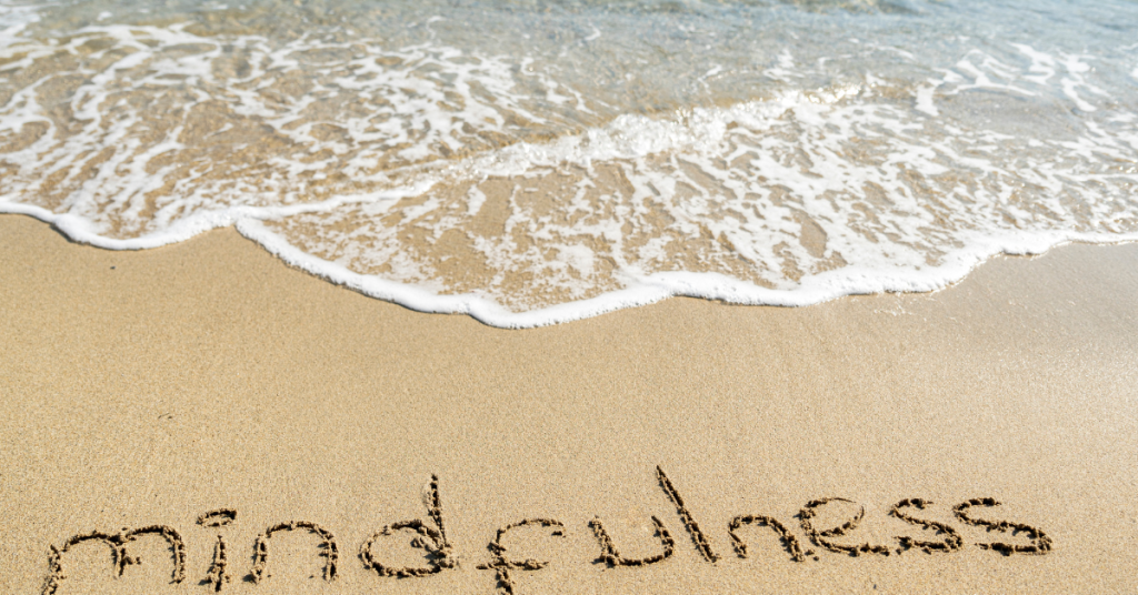 ocean waves washing up on a beach with the word mindfulness spelt out in the sand.
